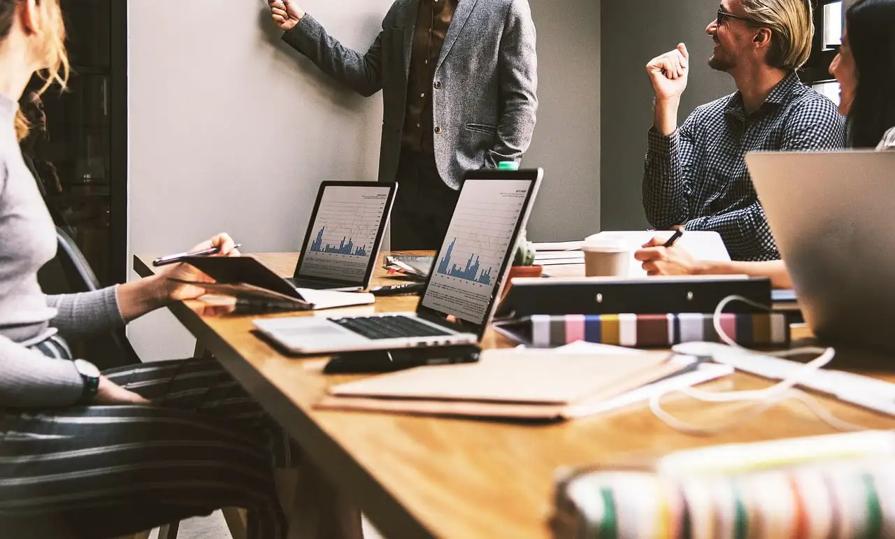 Business meeting with presenter at whiteboard, engaging participants seated around a table with laptops and technology, focusing on collaborative discussions for IT and digital marketing solutions.